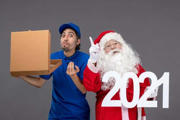 Vista frontal de santa claus con mensajero masculino sosteniendo bolsas de compras y caja de comida en la pared gris