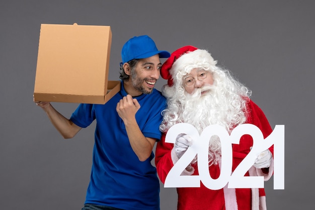 Vista frontal de santa claus con mensajero masculino sosteniendo bolsas de compras y caja de comida en la pared gris