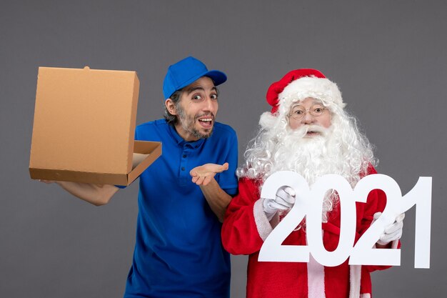 Vista frontal de santa claus con mensajero masculino sosteniendo bolsas de compras y caja de comida en la pared gris