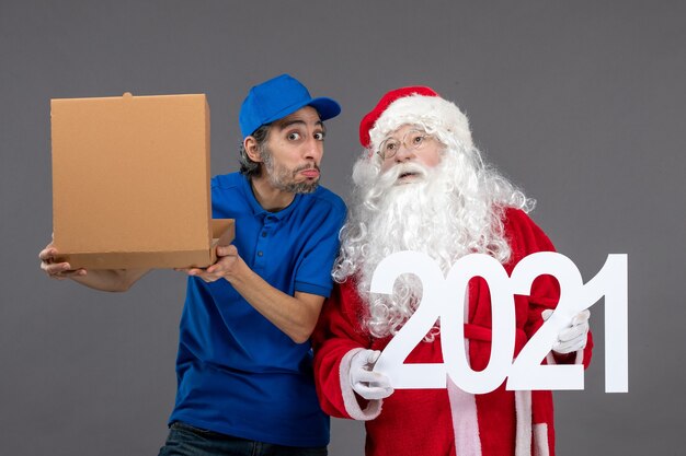 Vista frontal de santa claus con mensajero masculino sosteniendo caja de comida y escribiendo en la pared gris