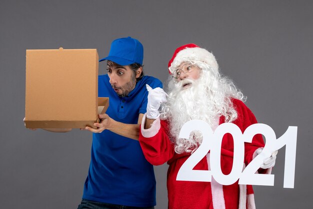 Vista frontal de santa claus con mensajero masculino sosteniendo caja de comida y escribiendo en la pared gris