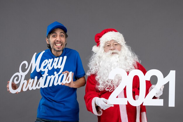 Vista frontal de santa claus con mensajero masculino sosteniendo feliz navidad y 2021 tableros en la pared gris