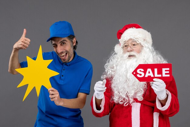 Vista frontal de santa claus con mensajero masculino sosteniendo pancarta de venta y cartel amarillo en la pared gris