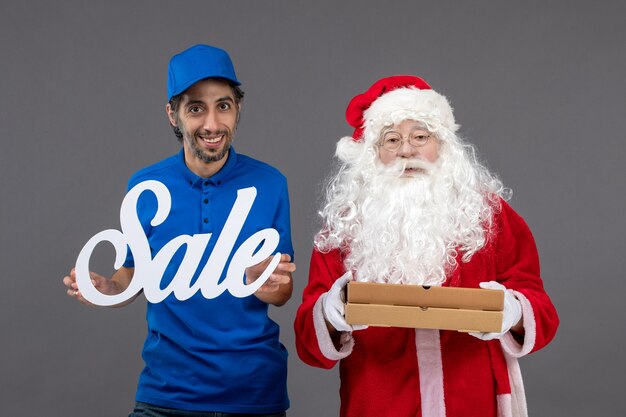 Vista frontal de santa claus con mensajero masculino sosteniendo pancartas de venta y cajas de alimentos en la pared gris