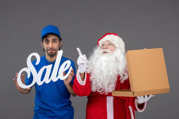 Vista frontal de santa claus con mensajero masculino sosteniendo pancartas de venta y cajas de alimentos en la pared gris