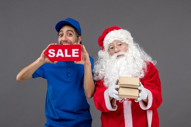 Vista frontal de santa claus con mensajero masculino sosteniendo pancartas de venta y paquetes de alimentos en la pared gris