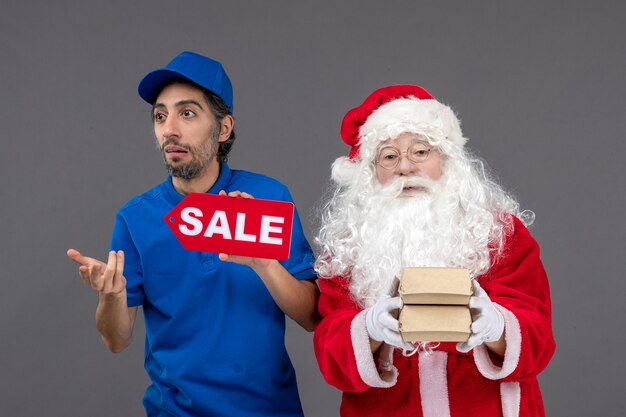 Vista frontal de santa claus con mensajero masculino sosteniendo pancartas de venta y paquetes de alimentos en la pared gris