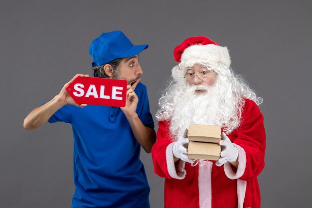 Vista frontal de santa claus con mensajero masculino sosteniendo pancartas de venta y paquetes de alimentos en la pared gris