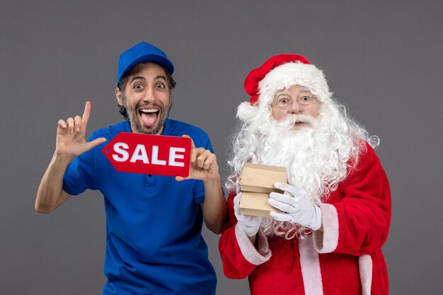 Vista frontal de santa claus con mensajero masculino sosteniendo pancartas de venta y paquetes de alimentos en la pared gris