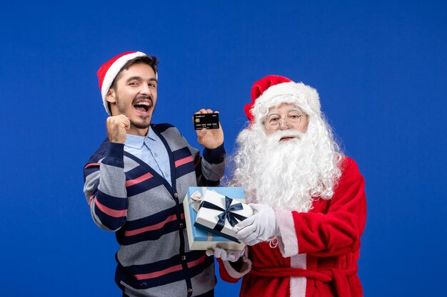 Vista frontal de santa claus con regalos y hombre joven con tarjeta bancaria en la emoción de Navidad azul