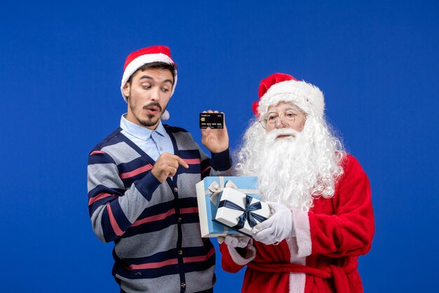Vista frontal de santa claus con regalos y hombre joven con tarjeta bancaria en las vacaciones de Navidad azul