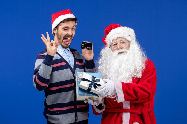 Vista frontal de santa claus con regalos y varones jóvenes con tarjeta bancaria en el escritorio azul vacaciones de navidad