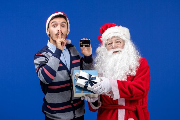 Vista frontal de santa claus con regalos y varones jóvenes con tarjeta bancaria en el piso azul vacaciones de navidad