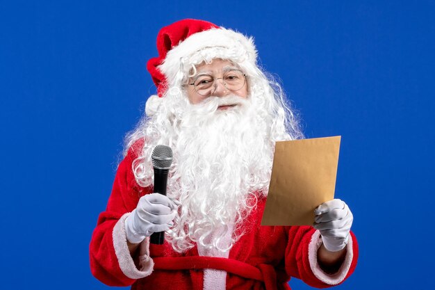 Vista frontal de santa claus sosteniendo el micrófono y leyendo la letra en el escritorio azul año nuevo color vacaciones nieve de navidad