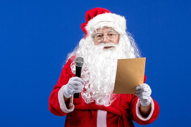 Vista frontal de santa claus sosteniendo el micrófono y leyendo la letra en la nieve de navidad de color azul año nuevo vacaciones