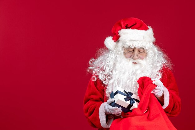 Vista frontal de santa claus sosteniendo el presente de la bolsa llena de regalos para niños en navidad de año nuevo rojo