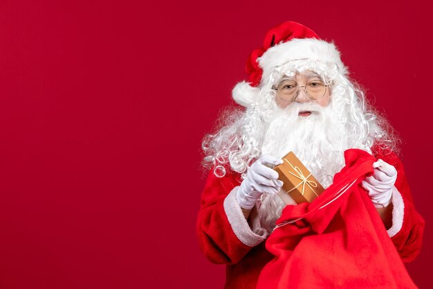 Vista frontal de santa claus sosteniendo el presente de la bolsa llena de regalos para niños en el piso rojo emoción año nuevo