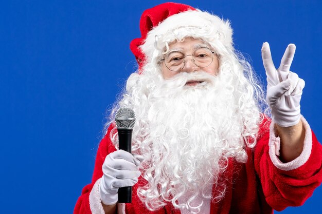 Vista frontal de santa claus con traje rojo y barba blanca sosteniendo el micrófono en el escritorio azul color vacaciones navidad año nuevo nieve