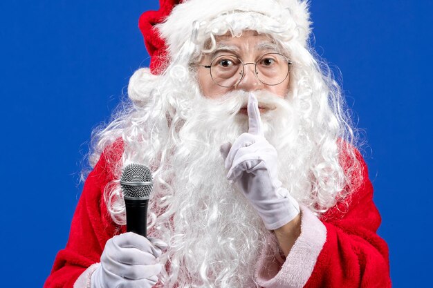 Vista frontal de santa claus con traje rojo y barba blanca sosteniendo el micrófono en una Navidad de vacaciones de color azul