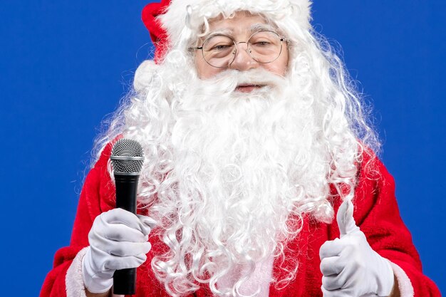Vista frontal de santa claus con traje rojo y barba blanca sosteniendo el micrófono en la nieve de año nuevo de navidad de vacaciones de color azul