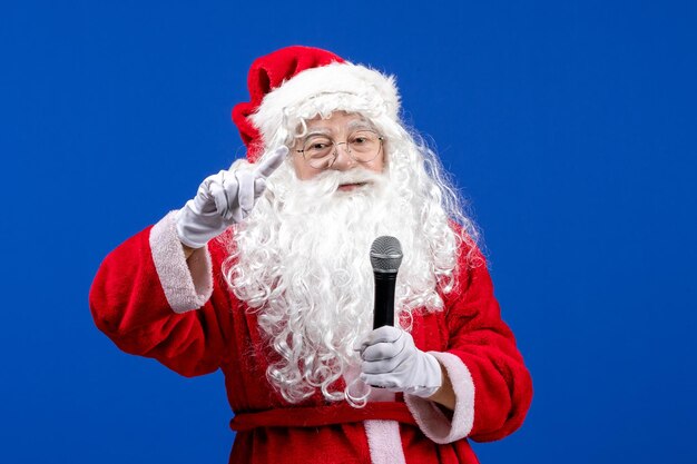 Vista frontal de santa claus con traje rojo y barba blanca sosteniendo el micrófono en la nieve azul vacaciones color de navidad año nuevo
