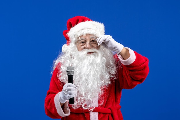 Vista frontal de santa claus con traje rojo y barba blanca sosteniendo el micrófono en la nieve de color azul vacaciones navidad año nuevo