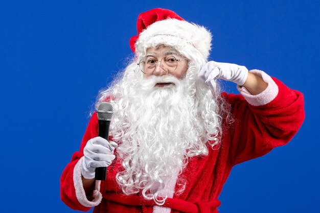 Vista frontal de santa claus con traje rojo y barba blanca sosteniendo el micrófono en vacaciones de color azul año nuevo