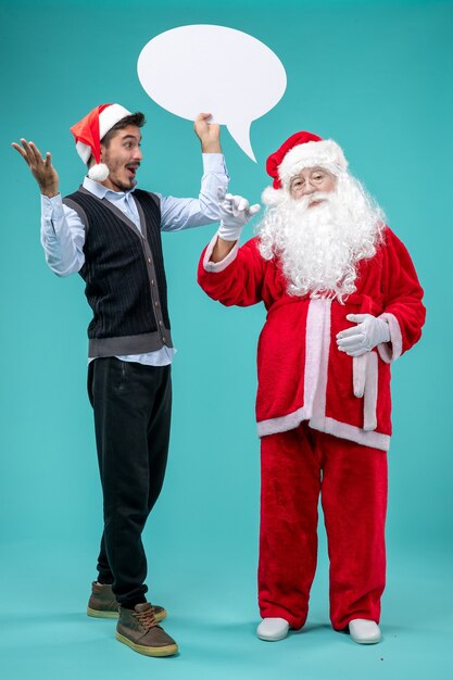 Vista frontal de santa claus con varones jóvenes que llevan a cabo un cartel blanco en el piso azul nieve navidad año nuevo color de vacaciones