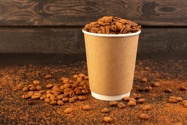 Una vista frontal de las semillas de café marrón dentro de un vaso de plástico sobre la superficie marrón y la semilla de café gránulo de grano oscuro