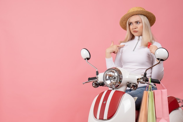 Vista frontal señorita en ciclomotor con bolsas de compras en pared rosa