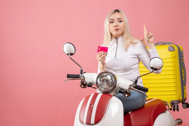 Vista frontal señorita en ciclomotor con tarjeta apuntando al techo elegante pared rosa