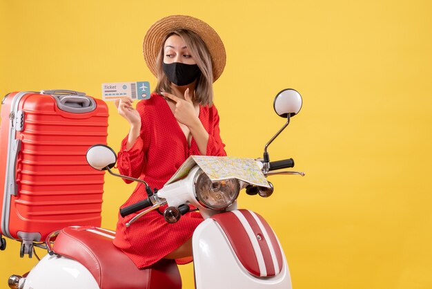 Vista frontal señorita con máscara negra en ciclomotor con maleta roja apuntando al billete