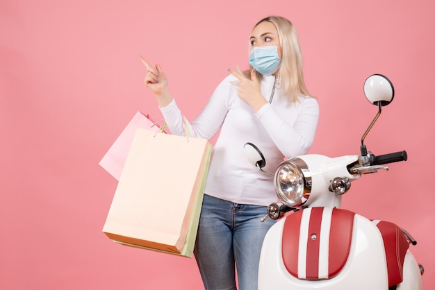 Vista frontal señorita sosteniendo bolsas de la compra apuntando con el dedo a la izquierda cerca del ciclomotor