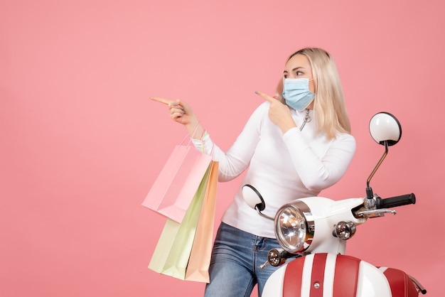 Vista frontal señorita sosteniendo bolsas de compras apuntando a la izquierda de pie cerca del ciclomotor