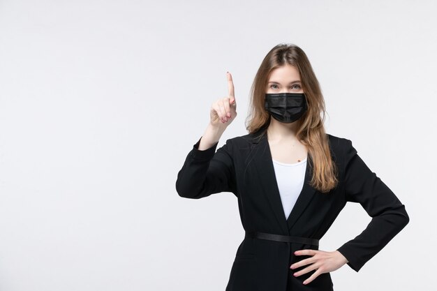 Vista frontal de la señorita en traje con mascarilla quirúrgica y apuntando hacia arriba en blanco