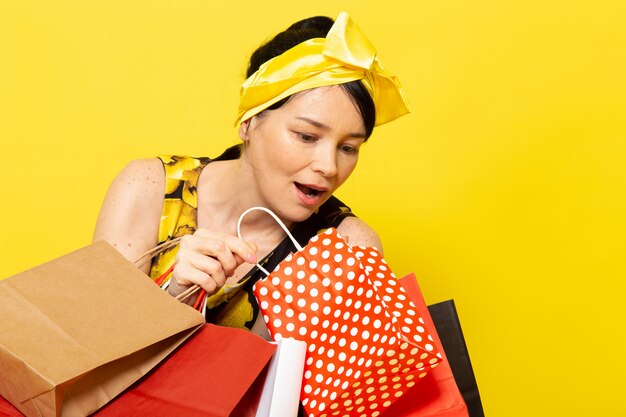 Una vista frontal señorita en vestido de flores amarillo-negro diseñado con vendaje amarillo en la cabeza posando sosteniendo paquetes de compras en el amarillo