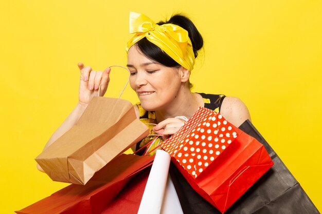 Una vista frontal señorita en vestido de flores amarillo-negro diseñado con vendaje amarillo en la cabeza posando sosteniendo paquetes de compras en el amarillo