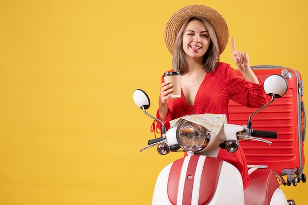 Vista frontal señorita en vestido rojo sacando la lengua sosteniendo la taza de café apuntando con el dedo hacia arriba