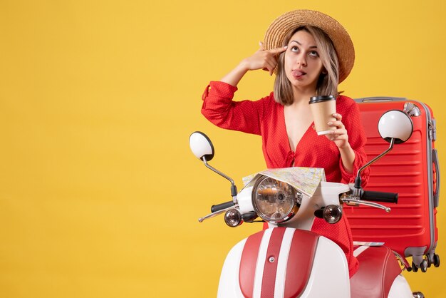 Vista frontal señorita en vestido rojo sacando la lengua sosteniendo la taza de café cerca del ciclomotor