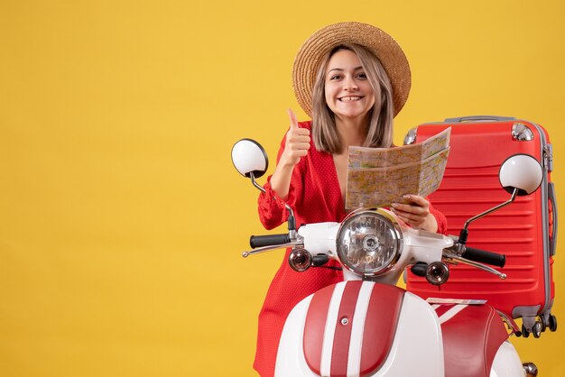 Vista frontal señorita en vestido rojo sosteniendo mapa dando pulgar hacia arriba cerca de ciclomotor