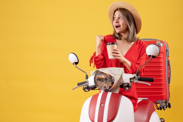 Vista frontal señorita en vestido rojo sosteniendo la taza de café apuntando detrás cerca del ciclomotor