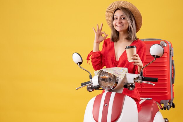 Vista frontal señorita en vestido rojo sosteniendo una taza de café gesticulando bien firmar cerca del ciclomotor