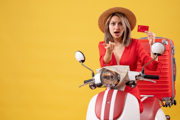 Vista frontal señorita en vestido rojo con tarjeta de crédito apuntando al ciclomotor cercano