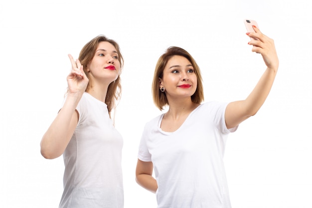 Una vista frontal señoritas en camisetas blancas tomando una selfie en el blanco