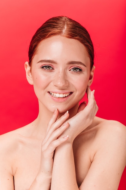 Vista frontal de la sensual chica caucásica con linda sonrisa. Foto de estudio de refinada mujer desnuda aislada sobre fondo rojo.