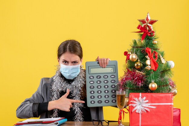 Vista frontal seria joven con máscara médica sentada en la mesa apuntada con calculadora de dedo árbol de navidad y cóctel de regalos