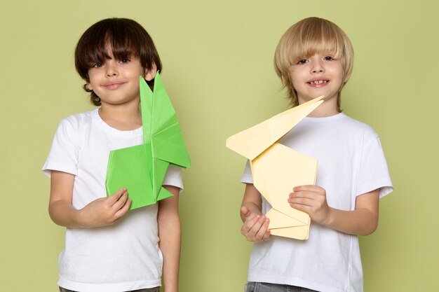 Una vista frontal sonriendo niños felices en camisetas blancas con coloridos juguetes de papel en el espacio de color piedra