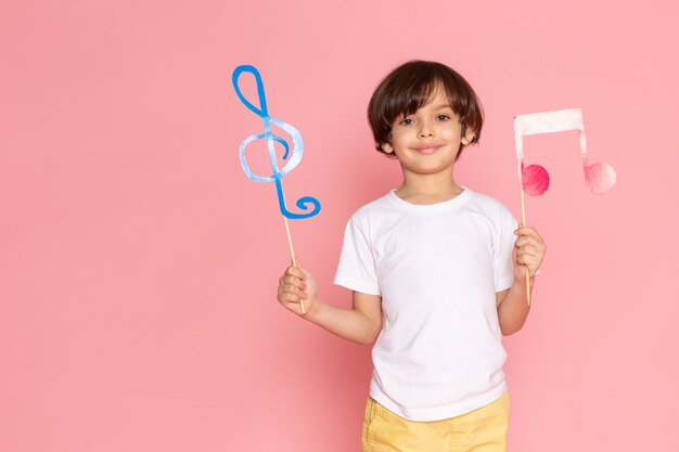 Una vista frontal sonriente adorable niño con carteles coloridos en camiseta blanca en el escritorio rosa