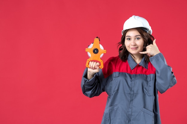 Vista frontal de la sonriente arquitecta en uniforme con casco sosteniendo cinta métrica y haciendo gesto de llamarme sobre fondo rojo aislado