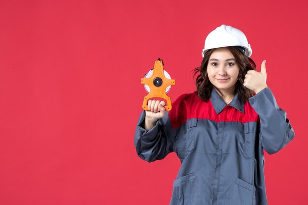 Vista frontal de la sonriente arquitecta en uniforme con casco sosteniendo cinta métrica y haciendo gesto ok sobre fondo rojo aislado